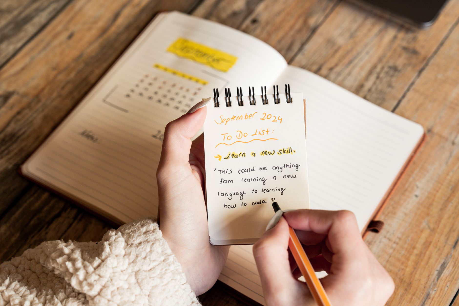 A person writes a specific learning goal in a paper notebook at a tidy desk with a pen and laptop nearby.