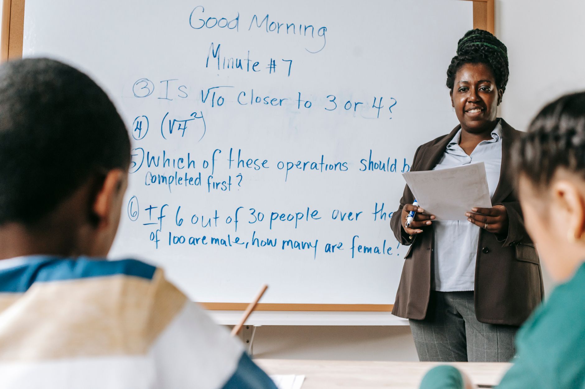 A teacher stands at a whiteboard and explains a concept with simple diagrams to a student.