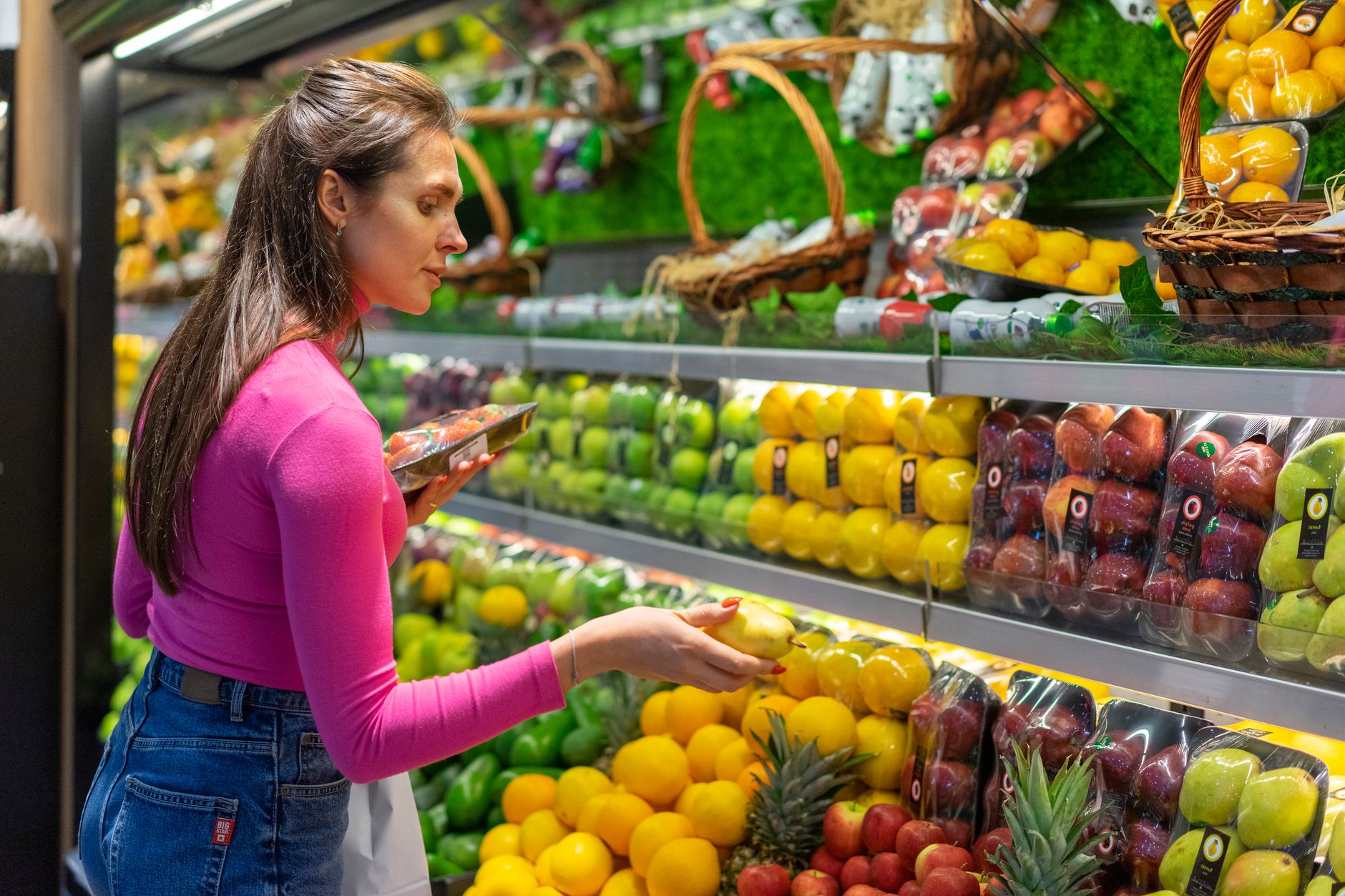 A person with a paper grocery list selecting vegetables in a supermarket produce aisle.