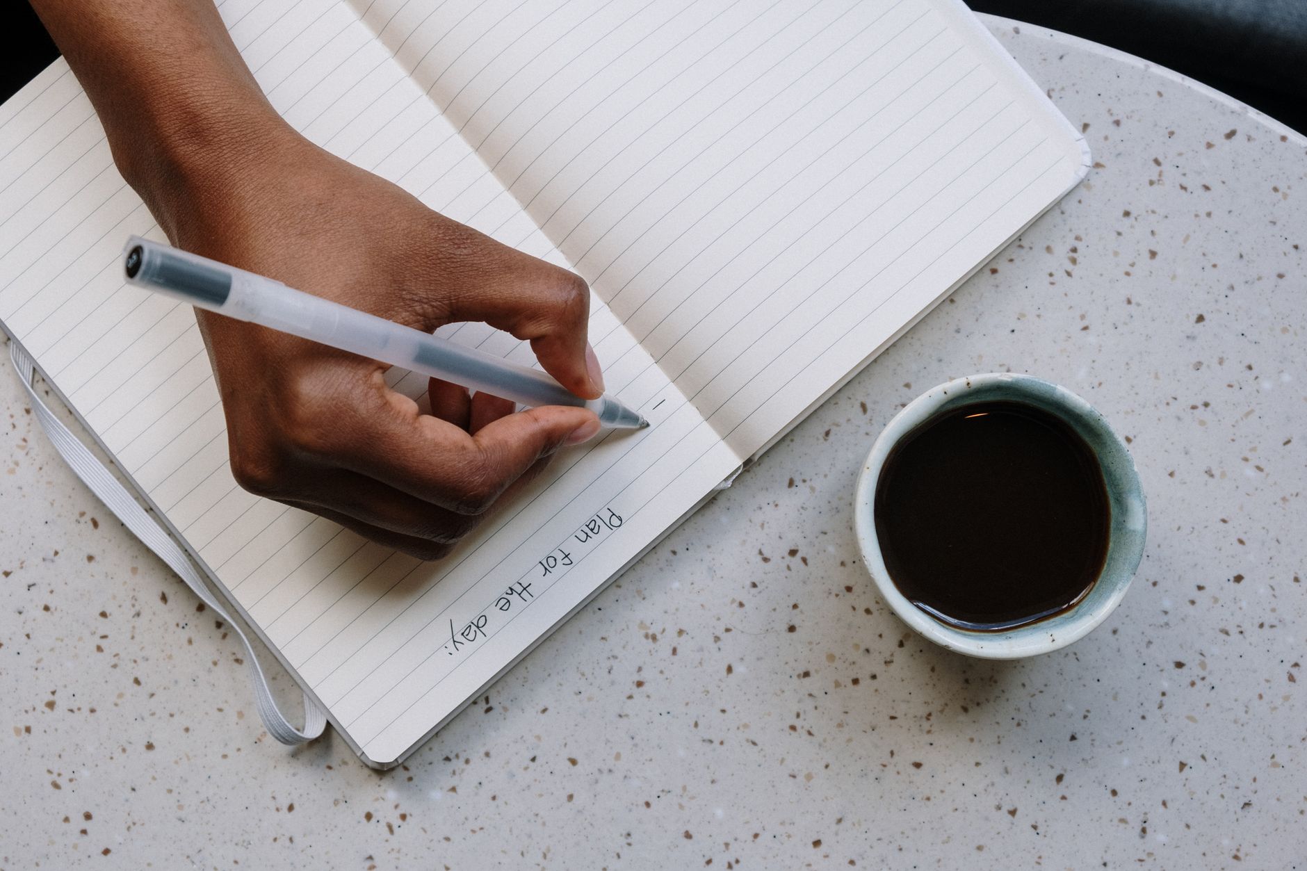 Person planning a weekly schedule in a paper planner with a coffee cup and phone on a wooden table.