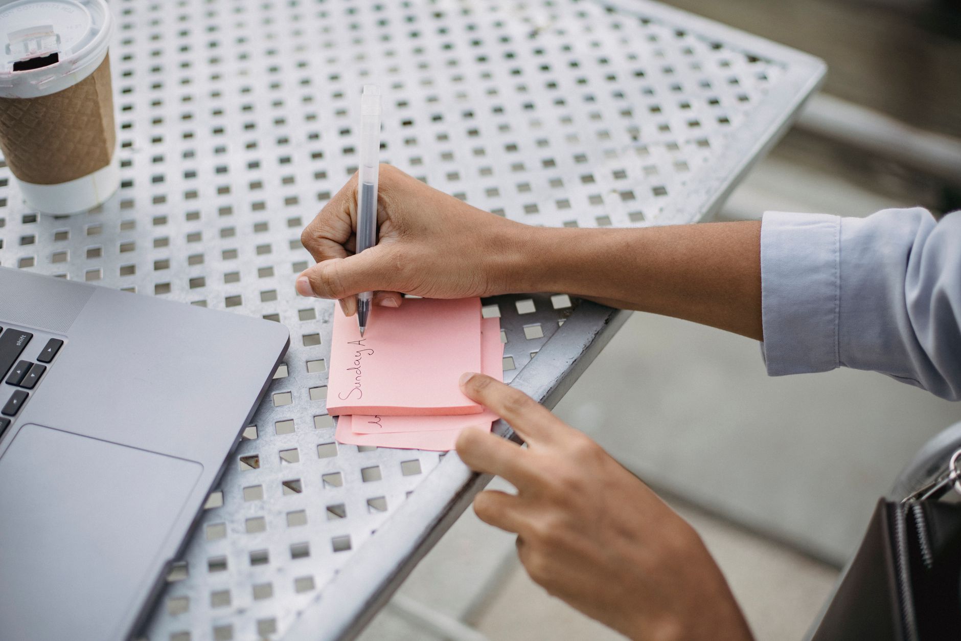 A person sits at a kitchen table writing a to-do list on paper next to a coffee mug and a smartphone.