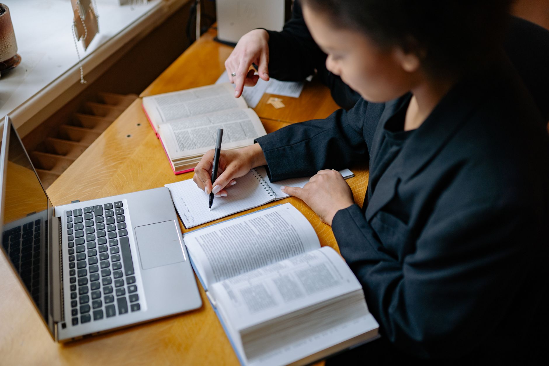 A student sits at a desk taking notes in a notebook next to an open laptop, with a mug and headphones nearby.