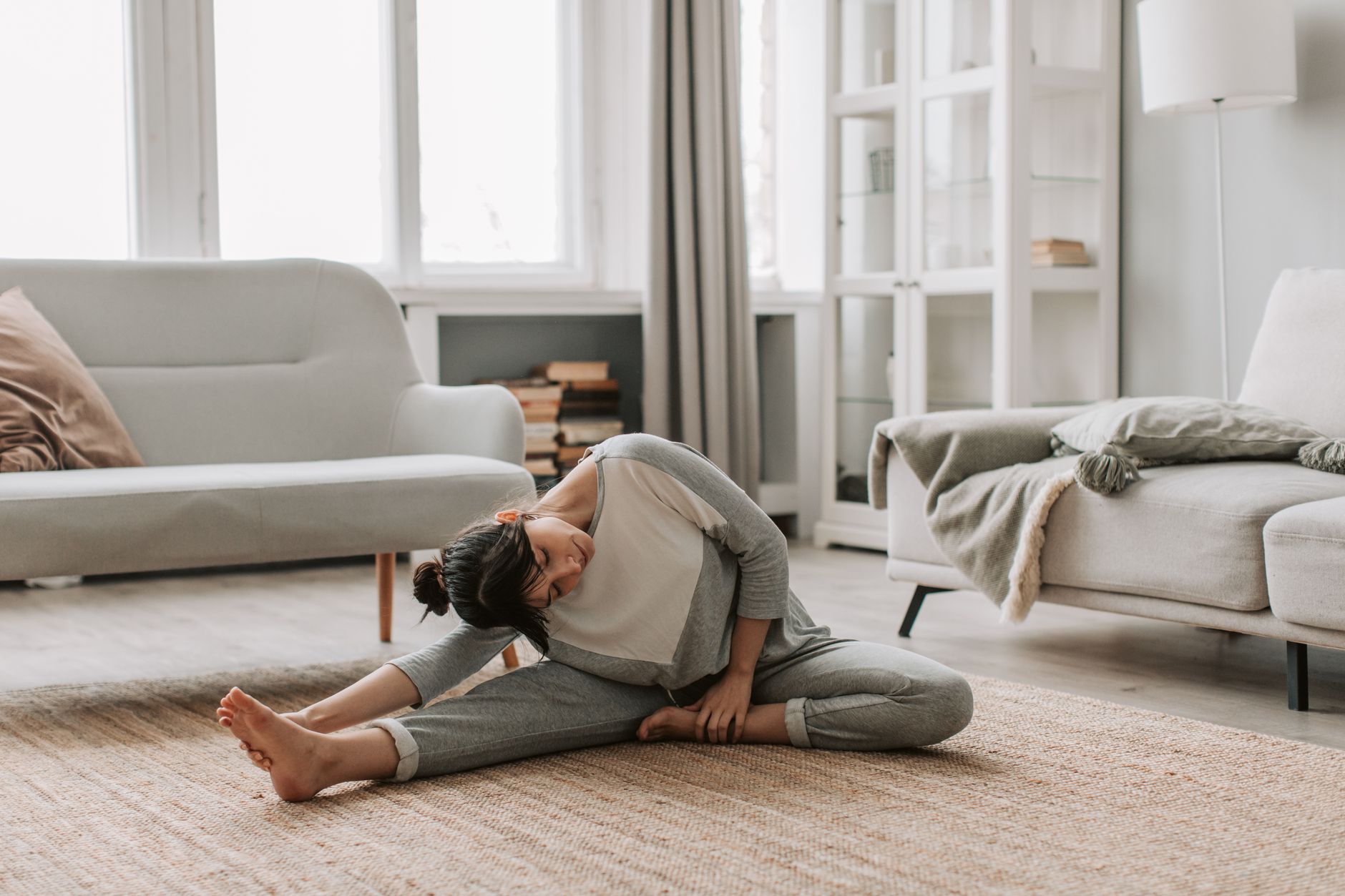 A person stretches on a yoga mat in a bright living room with natural light, wearing comfortable workout clothes.