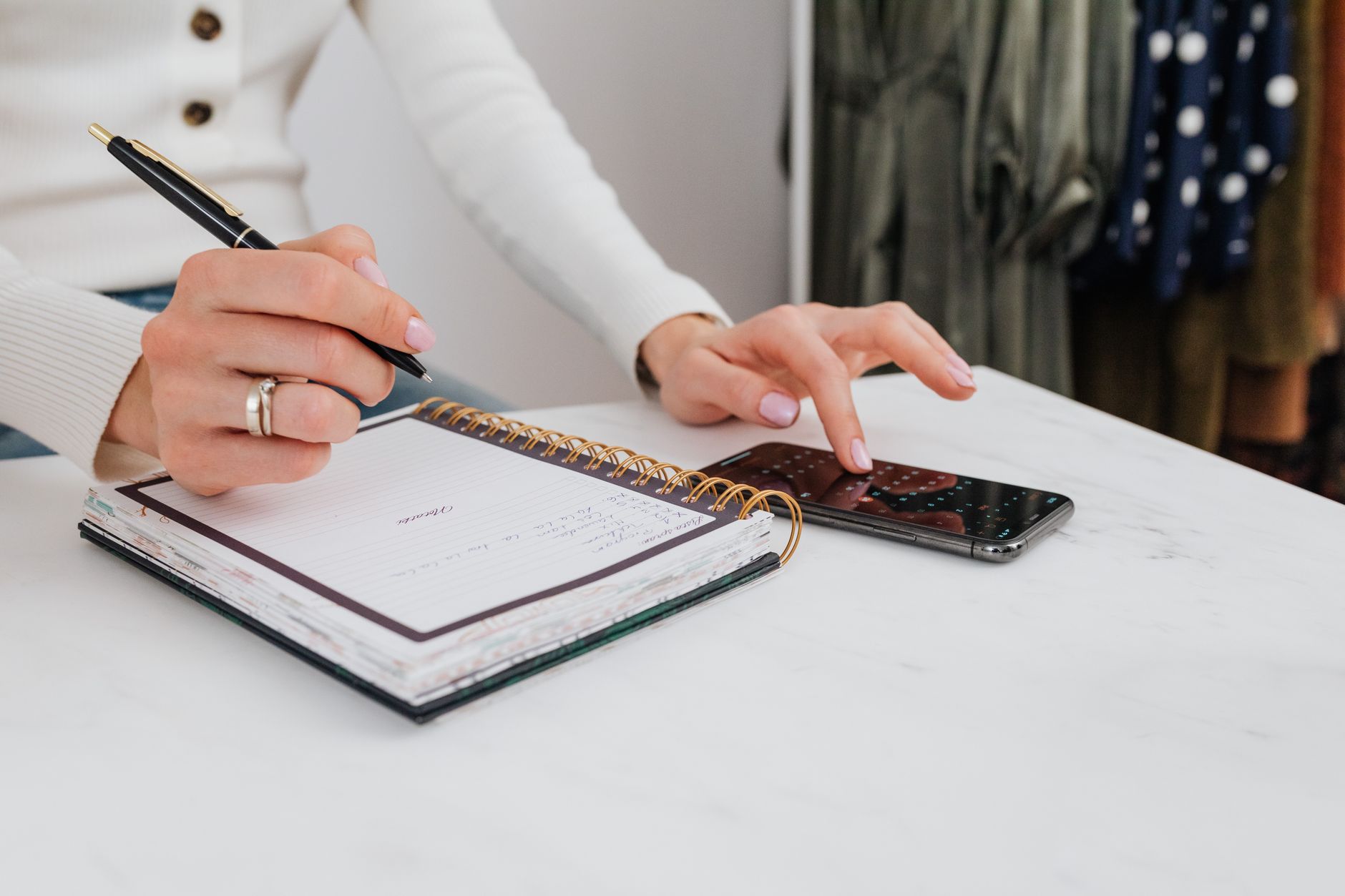 A person writes a daily schedule in a paper notebook while a smartphone sits on the table.