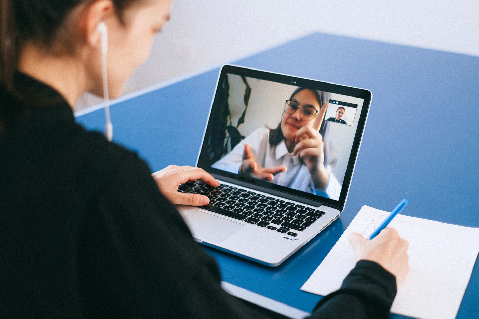 A student watches an online video on a laptop and takes handwritten notes in a notebook.